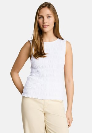 Młoda kobieta o długich, jasnobrązowych włosach, ubrana w bezrękawnik biały top z fakturą oraz beżowe spodnie, stojąca na tle jednolitego tła.