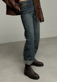 Personne portant un jean bleu foncé à revers, des bottes Chelsea en cuir marron et une veste en cuir marron avec une fermeture éclair.