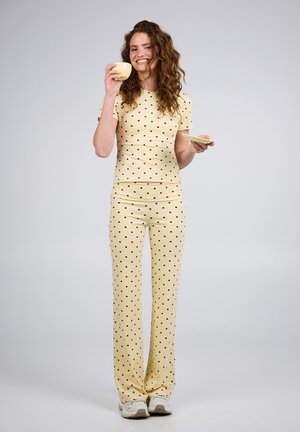 Smilende kvinne i gul polkadot-antrekk som holder en gul kopp og underkop, stående mot en ensfarget lys grå bakgrunn.