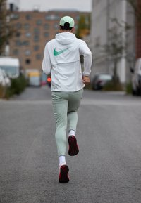 Ľahká biela bežecká bunda s zeleným logom Nike, mintové priliehajúce nohavice a bežecké topánky s čiernou podrážkou a červenými akcentmi.