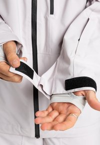 Hände, die einen weißen Jackenärmel mit schwarzem Klettverschluss, Daumen-Schlaufe am inneren Ärmel und teilweise sichtbarem Frontreißverschluss anpassen.