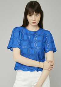 Haut en dentelle bleue avec un ourlet festonné, des découpes circulaires et des manches courtes. Le tissu a une texture lisse et des détails de couture décoratifs.