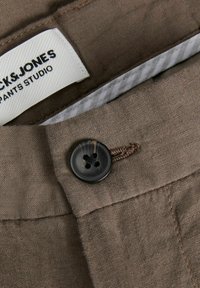 Καφέ ύφασμα με υφή, που διαθέτει ένα μαύρο κουμπί και μια λευκή ετικέτα που γράφει "Jack & Jones Pants Studio" σε μια γωνία.