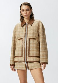 Chaqueta de cuadros marrón con un cuello de cuero marrón, dos grandes bolsillos y una cremallera central. La tela tiene un patrón de tejido texturizado.