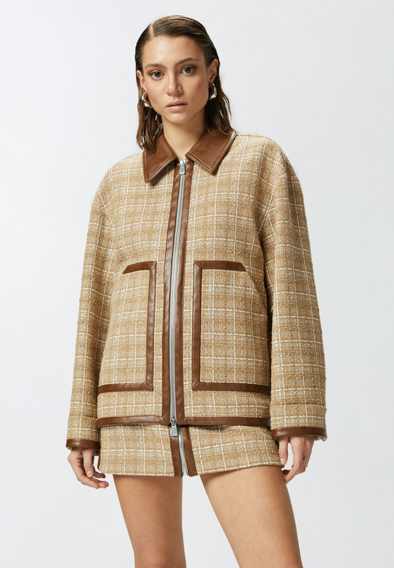 Chaqueta de cuadros marrón con un cuello de cuero marrón, dos grandes bolsillos y una cremallera central. La tela tiene un patrón de tejido texturizado.