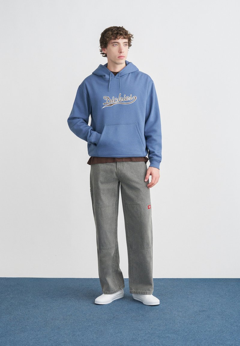 Felpa con cappuccio blu con logo bianco "Dickies", pantaloni a gamba larga grigi con etichetta rossa e scarpe da ginnastica bianche. Sfondo grigio chiaro.