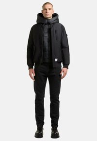 Schwarze Steppjacke mit Kapuze über einer gesteppte schwarze Weste, kombiniert mit schwarzen Jeans und schwarzen Stiefeln, mit Reißverschluss und Taschen.