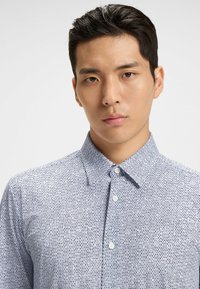 Blå mönstrad skjorta med knappar och en strukturerad krage; har ett geometriskt mönster i mörkblått och vitt; slät tygtextur.