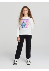 Biała koszula z długim rękawem z kolorową grafiką kreskówkową przedstawiającą dwie postacie, zestawiona z czarnymi dresami i czarno-białymi trampkami.