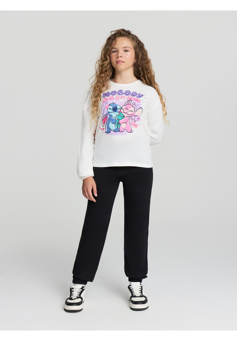 Biała koszula z długim rękawem z kolorową grafiką kreskówkową przedstawiającą dwie postacie, zestawiona z czarnymi dresami i czarno-białymi trampkami.