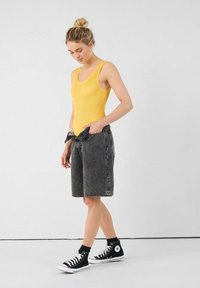 Top sin mangas amarillo acanalado combinado con shorts de mezclilla oscuros desgastados y zapatillas negras de caña alta, destacando un look casual pero contemporáneo.