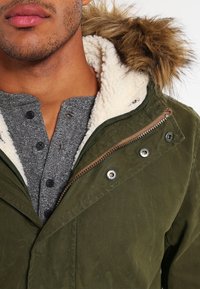 Grön jacka med pälsfodrad huva, med dragkedja och tryckknappar, samt en grå strukturerad henley-tröja under.