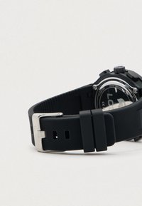 Μαύρο ψηφιακό ρολόι με υφασμάτινο λουρί από καουτσούκ και μεταλλικό κούμπωμα, παρουσιαζόμενο από την πίσω πλευρά σε άσπρο φόντο.