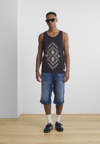 Black tank top with white geometric pattern, denim shorts, white socks, and black sneakers. Plain background with wooden floor.