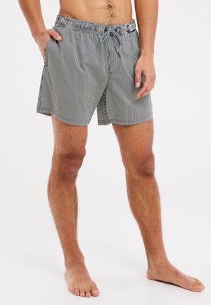 Man draagt zwart-witte zwemshorts met verticale strepen en een trekkoord, staat op blote voeten met één hand in de zak tegen een witte achtergrond.