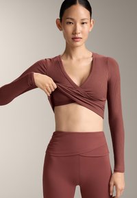 Donna che solleva una maglia a maniche lunghe stile incrociato color bordeaux per mostrare reggiseno sportivo abbinato e leggings a vita alta su sfondo beige.