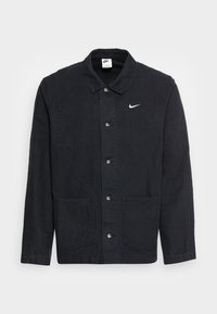 Svart bomullsjacka med krage, knappstängning, två stora framfickor och en liten vit Nike-logotyp på bröstet.