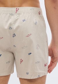 Μπέζ σορτς με ναυτικό σχέδιο που περιλαμβάνει βάρκες, ψάρια, αστακούς, πυξίδες και σημαίες, φορεμένα από ένα άτομο που φαίνεται από τη μέση και κάτω.