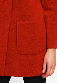 Punainen villakangastakki, jossa on teksturoitu pinta, pyöreä tasku edessä ja näkyvät saumat reunoilla.