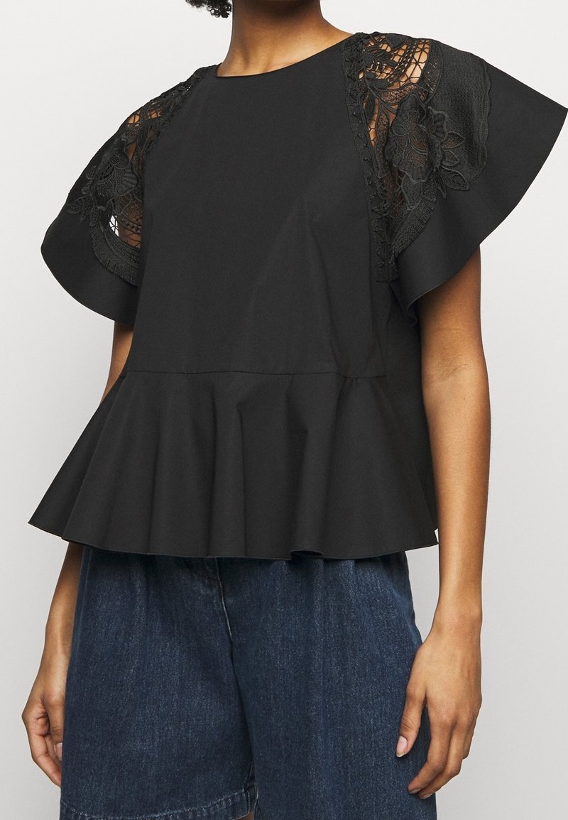 Femme portant un chemisier noir à manches courtes avec des détails en dentelle aux épaules et un ourlet à volants, combiné à un short en jean bleu foncé.