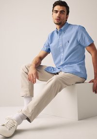 Šviesiai mėlyna trumpasleeve marškinėliai su mažai siuvinėtu logotipu. Derinami su smėlinėmis kelnėmis ir baltomis snukiais, sėdintys neutraliame fone.