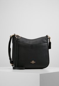 Black leather shoulder bag with gold hardware, adjustable strap, and front logo emblem, displayed on white pedestal against a neutral background.