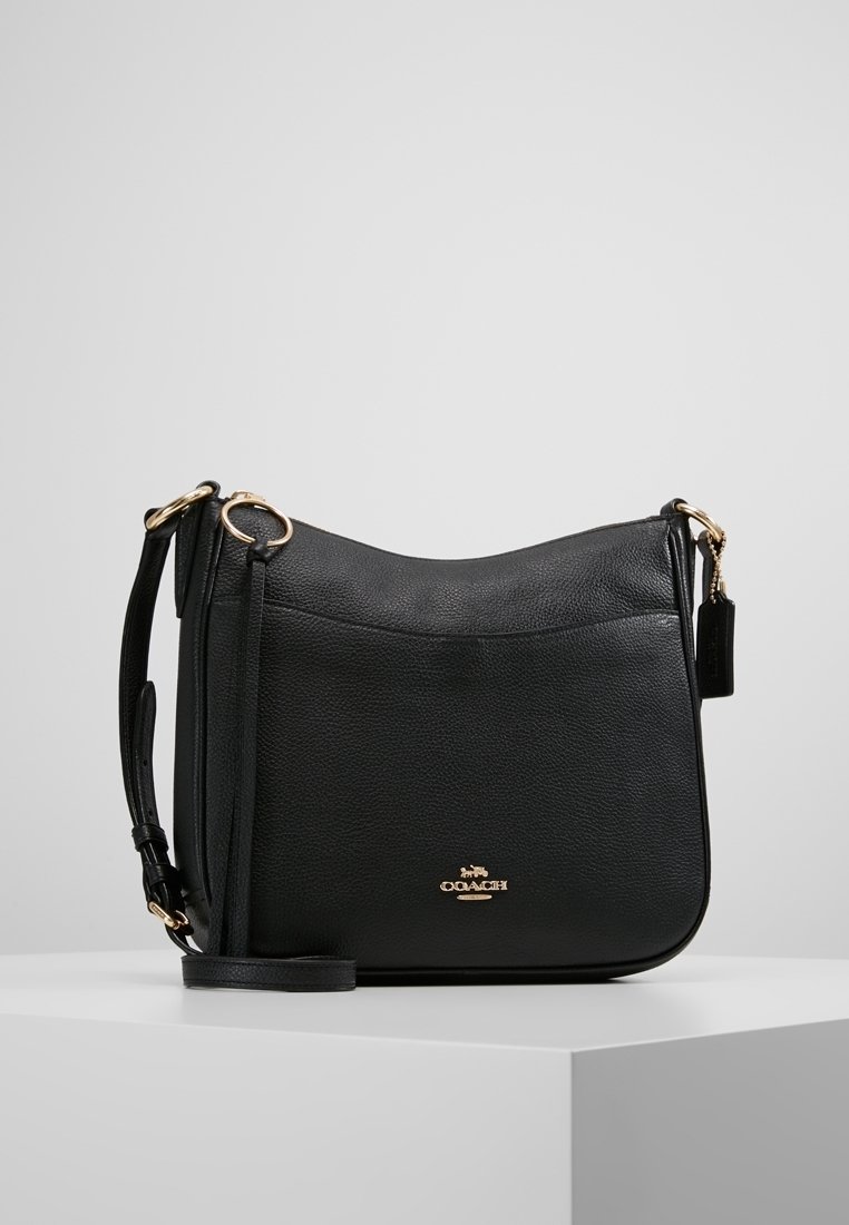 Black leather shoulder bag with gold hardware, adjustable strap, and front logo emblem, displayed on white pedestal against a neutral background.