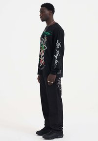 Zwarte langarmshirt met kleurrijke grafische ontwerpen en witte tekst. Gecombineerd met zwarte broek met een spinnenwebpatroon aan één kant.