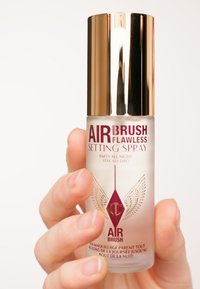 Clear glass bottle with a gold cap; red text. Label reads "AIRBRUSH FLAWLESS SETTING SPRAY." Features an emblem with wings.