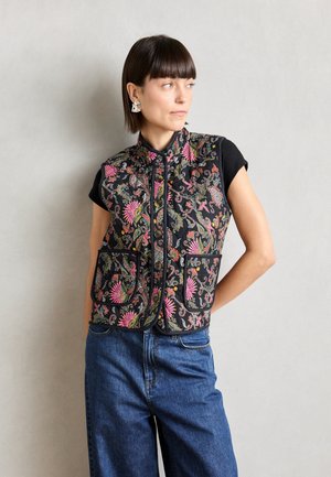 Chaleco negro con bordado floral colorido, cuello alto, botones delanteros y dos bolsillos. Lleva puesto encima de una camiseta de manga corta negra y jeans.