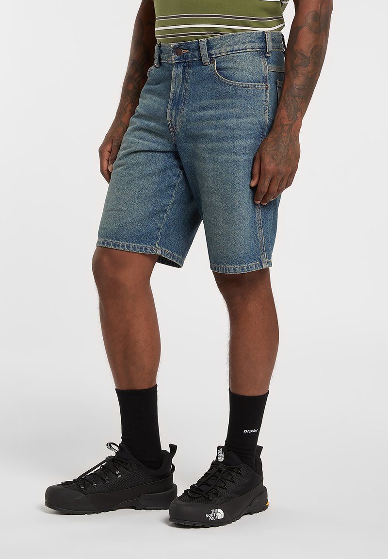 Uomo con pantaloncini di jeans blu, calzini neri al polpaccio e scarpe nere The North Face, in piedi contro uno sfondo bianco.