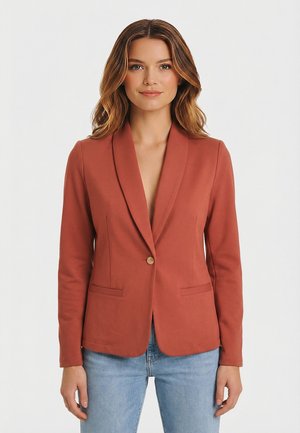 Vrouw draagt een roestkleurig colbert met een enkele knoop en klepzakken, gecombineerd met lichtblauwe spijkerbroek, staand tegen een effen witte achtergrond.