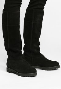 Černé semišové kozačky po kolena s plochou gumovou podrážkou, vyznačující se hladkým povrchem a jednoduchým prošíváním, navržené pro těsné padnutí.