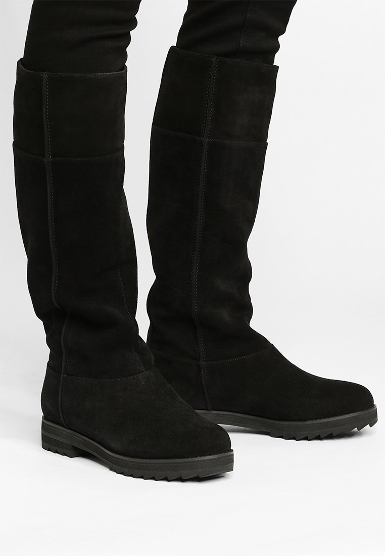 Černé semišové kozačky po kolena s plochou gumovou podrážkou, vyznačující se hladkým povrchem a jednoduchým prošíváním, navržené pro těsné padnutí.