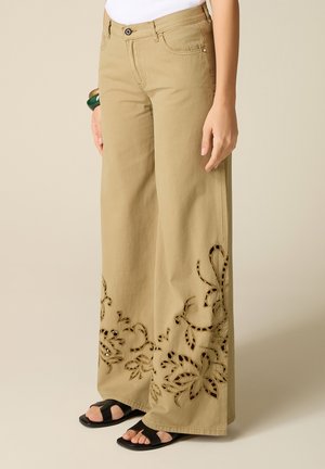 Pantaloni beige a gamba larga con ricamo floreale traforato sull'orlo, indossati con sandali neri open toe e bracciali verdi e marroni.