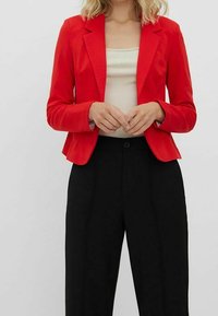 Blazer rouge à coupe ajustée, col à revers et manches longues. Porté sur un top crème, associé à un pantalon noir taille haute.