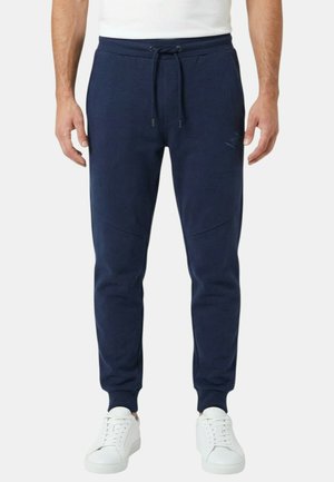 Homme portant un pantalon de jogging bleu marine avec taille à cordon et des baskets blanches, debout devant un fond uni.