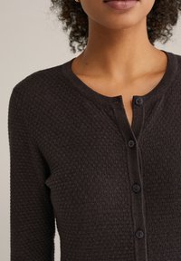 Temno rjav teksturiran kardigan z okroglim izrezom, s petimi črnimi gumbi in rahlo odprto zaponko.