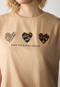 Beige t-shirt med tre hjerteformer i forskellige dyreprintmønstre: leopard, zebra og leopard igen, med sort tekst nedenfor.