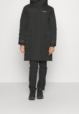 Persoon die een zwarte knielange parka met klepzakken, zwarte broek en zwarte wandelschoenen draagt, staand tegen een effen achtergrond.