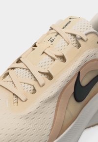 Μπεζ αθλητικό παπούτσι Nike με ύφασμα δίχτυ, κρέμ κορδόνια και μαύρο λογότυπο Nike στη πλευρά, κοντινή λήψη.