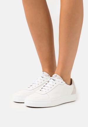 Piernas que llevan zapatillas blancas de cuero con cordones y detalle en el talón de ante beige, mostradas sobre un fondo blanco liso.