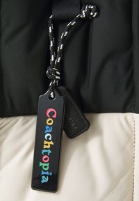 Căptușeală de jachetă neagră și bej, cu o etichetă neagră ce conține text colorat "Coachopia" și un detaliu din sârmă împletită neagră. Accente hardware simple.