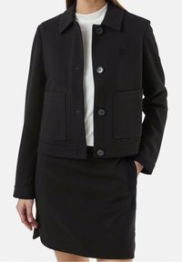 Czarna teksturowana kurtka z kołnierzykiem, zapinana na guziki z przodu, z dwiema dużymi kieszeniami. Nosi zakładkę na białej bluzce i czarnej spódnicy.