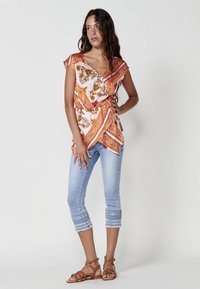 Blusa de estampado floral en blanco, naranja y verde; combinada con unos pantalones capri azul claro con puños bordados y sandalias marrones de tiras.