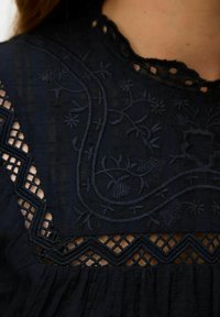 Mörkblå skjorta med texturerad tyg, blommig broderi och spetsdetaljer runt halsringningen och bröstområdet.