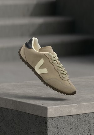Beige suede sneaker with cream "V" logo, laces, black heel tab, and textured brown sole floating above a gray concrete ledge.