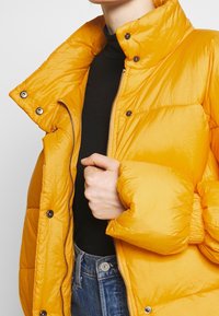 Giacca imbottita gialla con colletto alto, chiusura con zip e bottoni a pressione. Presenta una texture lucida e una vestibilità leggermente oversize.