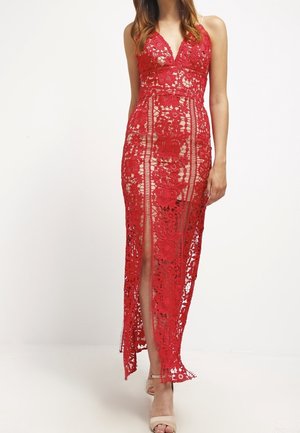 Kvinde iført en ærmeløs rød maxi-kjole i blonder med blomsterprint og en slids foran, kombineret med beige åbne hæle.