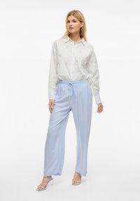 Camicia bianca a maniche lunghe con intricate pattern blu, abbinata a pantaloni leggermente blu e larghi e sandali con tacco trasparente.